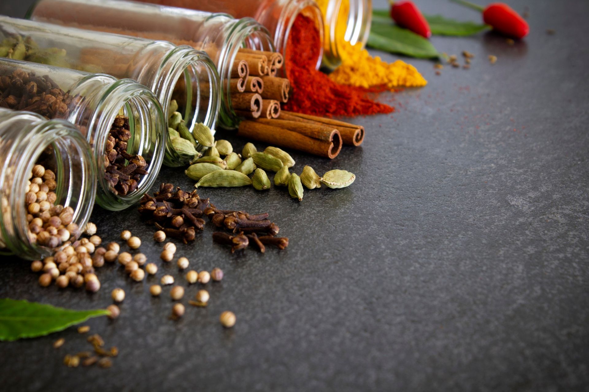 Spices spilling from jars: coriander seeds, cardamom, cinnamon sticks, chili peppers, and turmeric on a dark surface.