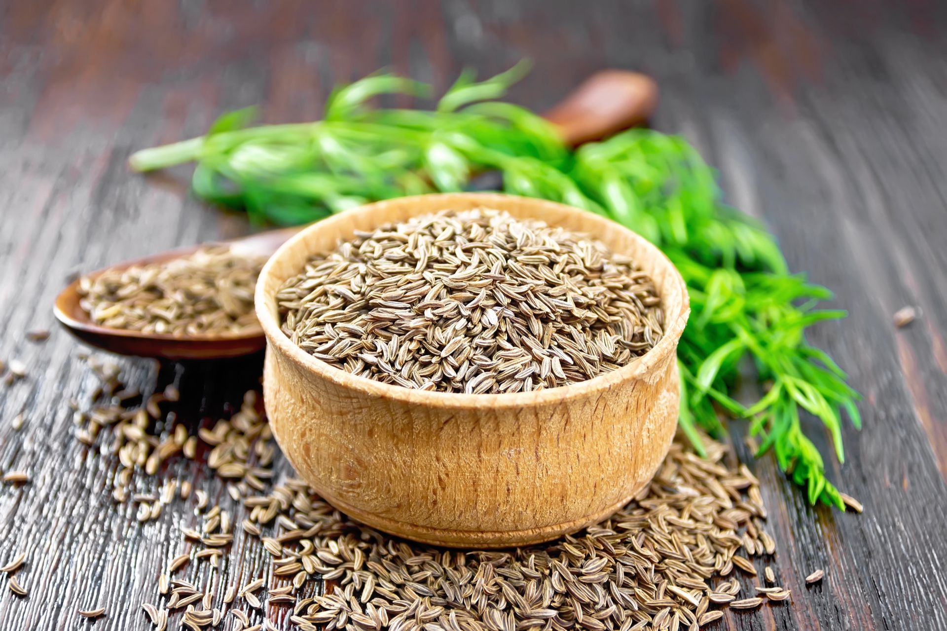 Wooden bowl filled with caraway seeds, with seeds spilled on a wooden surface, green herbs in the background.