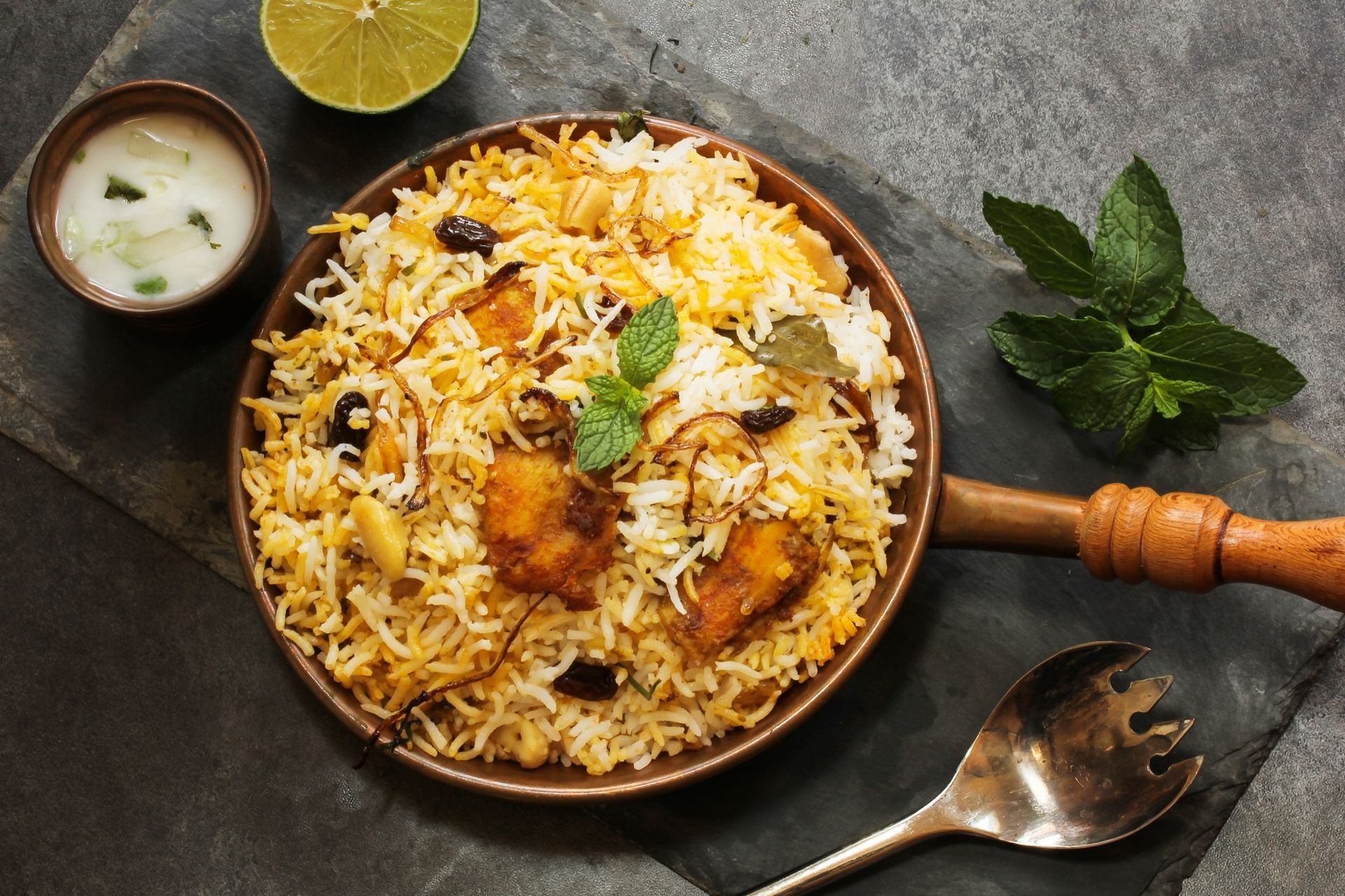 Chicken biryani with raita, lemon slice, and mint leaves on a dark surface.