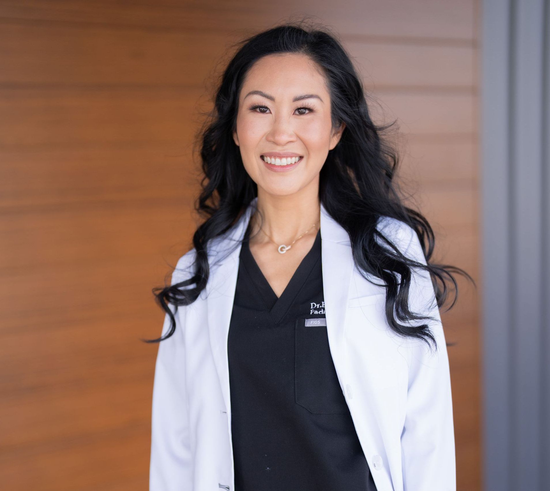 A woman in a white coat and black scrubs is smiling.