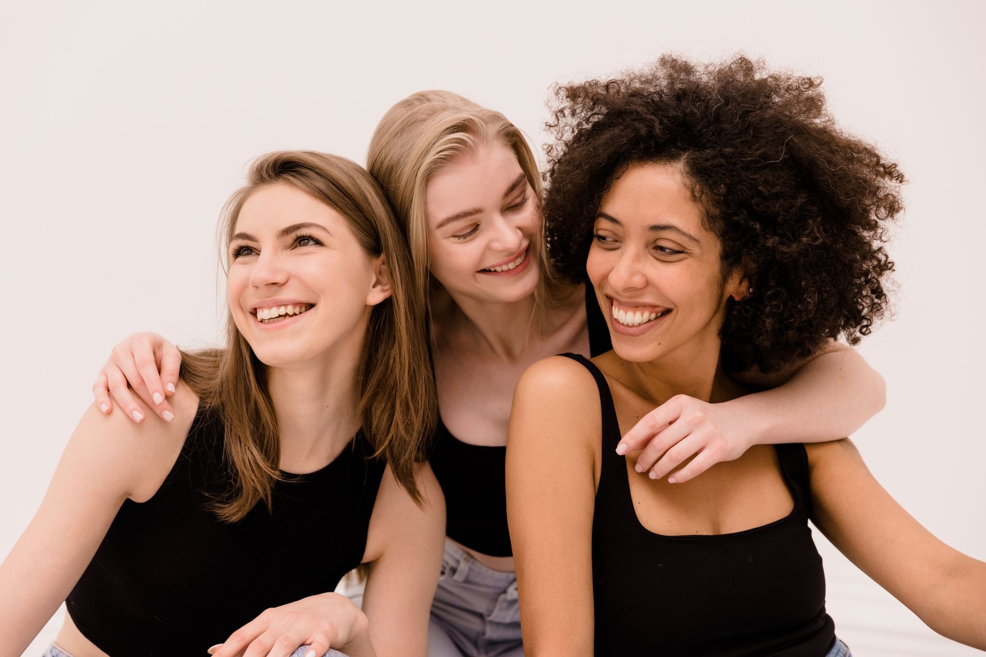 Three women are hugging each other and smiling while sitting on a bed.