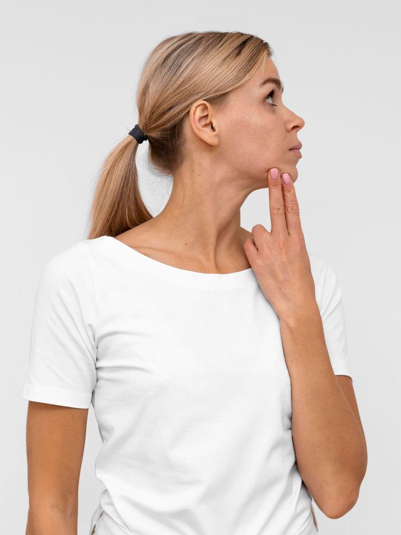 A woman in a white t-shirt is holding her hand to her chin.