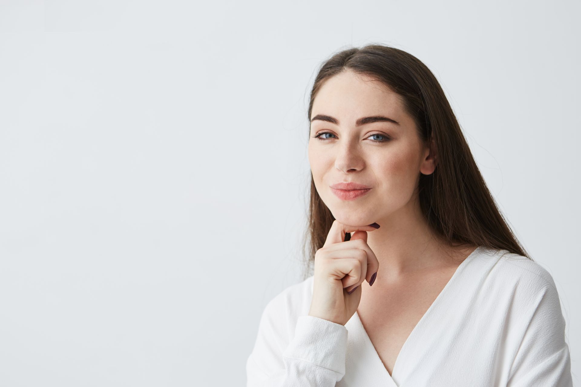 Woman in white shirt with hand under chin, looking thoughtful, against white background.