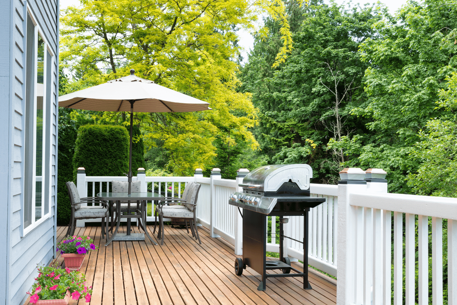 A wooden deck with a grill, a table and chairs under an umbrella, and surrounding trees in a backyard.