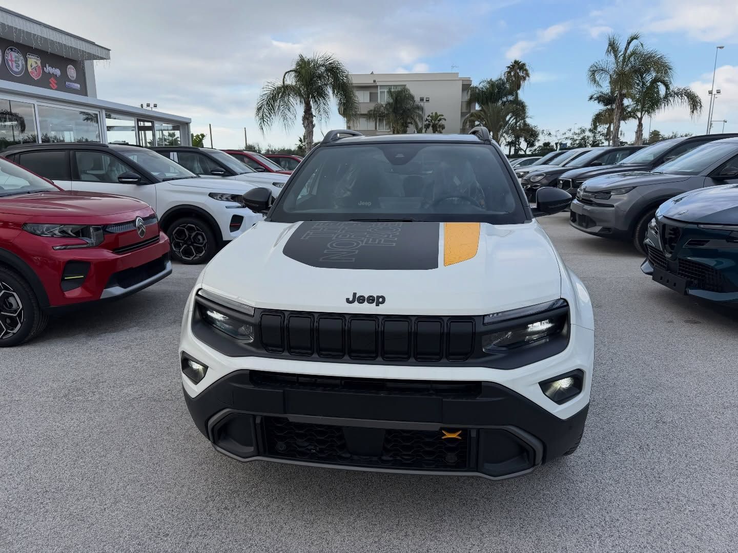 Un SUV Jeep bianco con dettagli neri e una striscia gialla sul cofano, parcheggiato tra altri veicoli.
