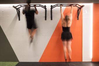Two people doing pull-ups on wall-mounted bars in a gym with orange and white accents.