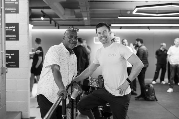 Two men smiling, leaning on railing, in a gym or office setting, other people in background.
