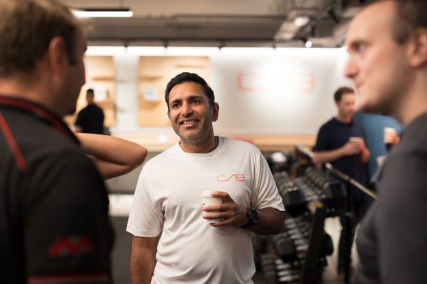 Three men talking in a gym, one holding a cup. Blurred weights and people in the background.