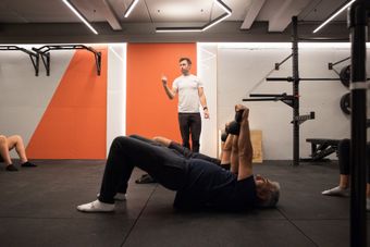 Man doing kettlebell exercise, coach instructing in a gym with orange wall and equipment.