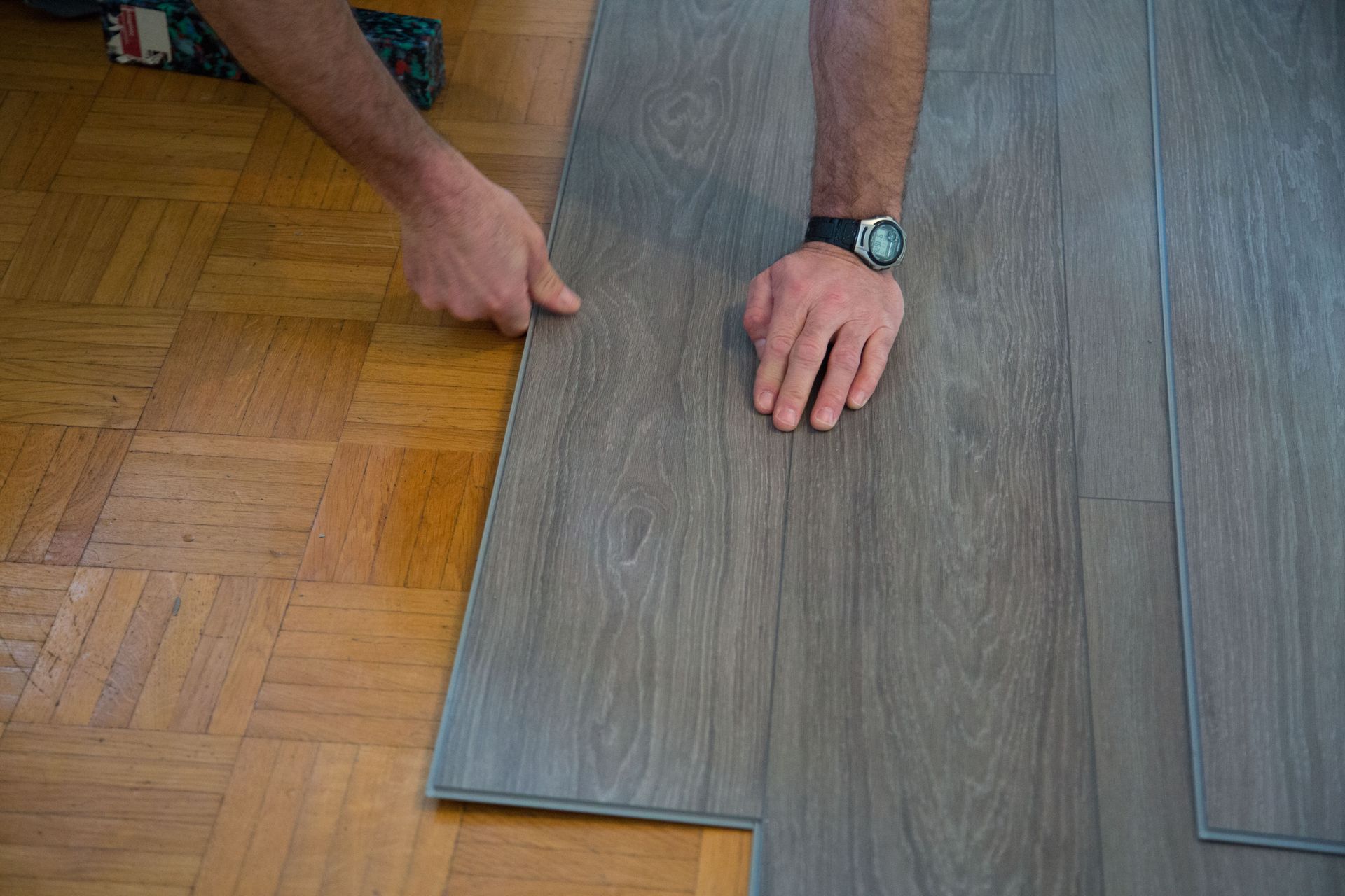 Hands pressing a gray wood-look laminate plank into place over existing parquet flooring.