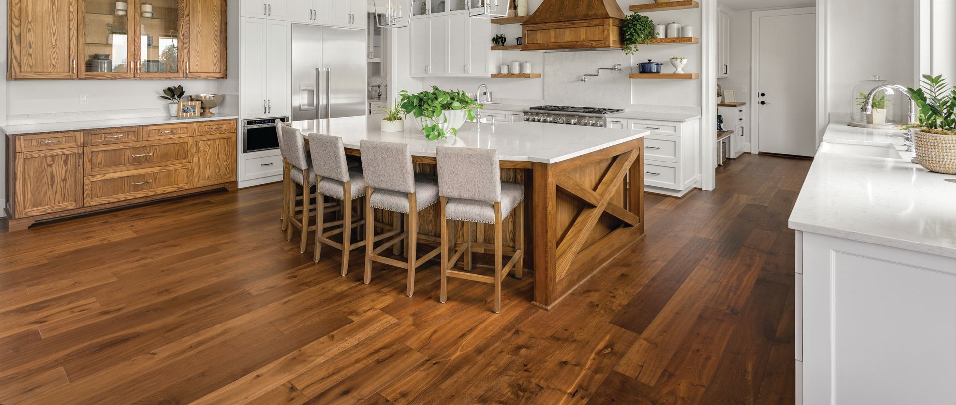 A modern kitchen with a large wooden island, white countertops, tan bar stools, and polished dark wood flooring.