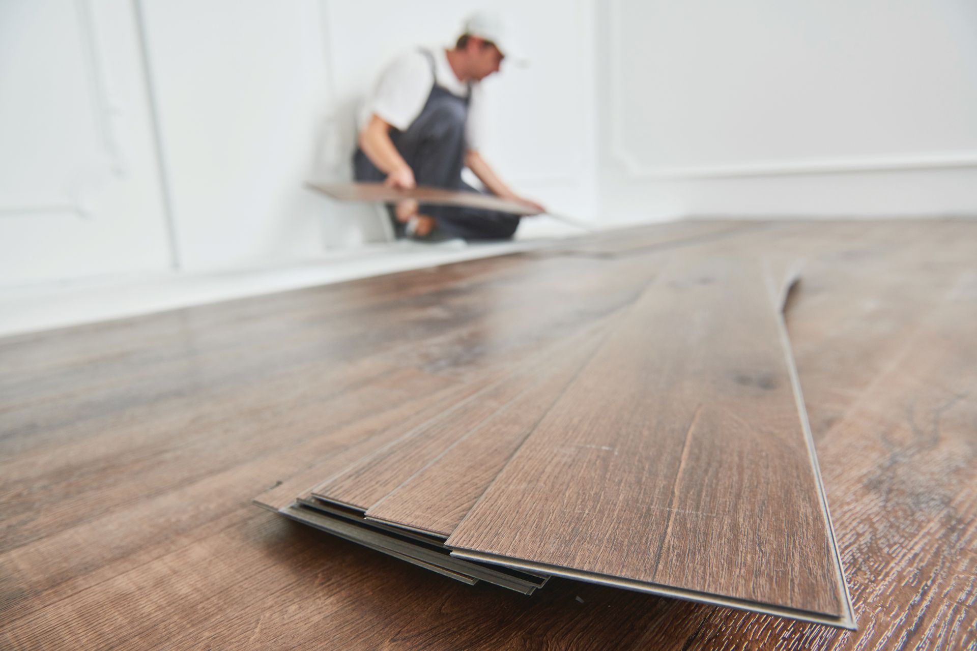 A construction worker installing brown, wood-patterned vinyl flooring planks in a room with white walls.
