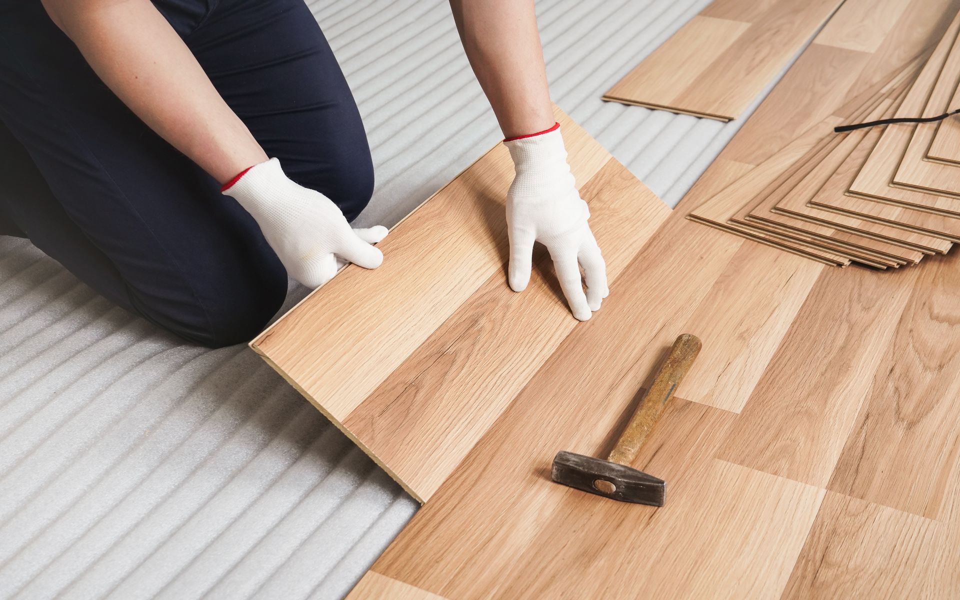 A person in white gloves installs wood-look laminate flooring on a grooved underlayment with a hammer nearby.