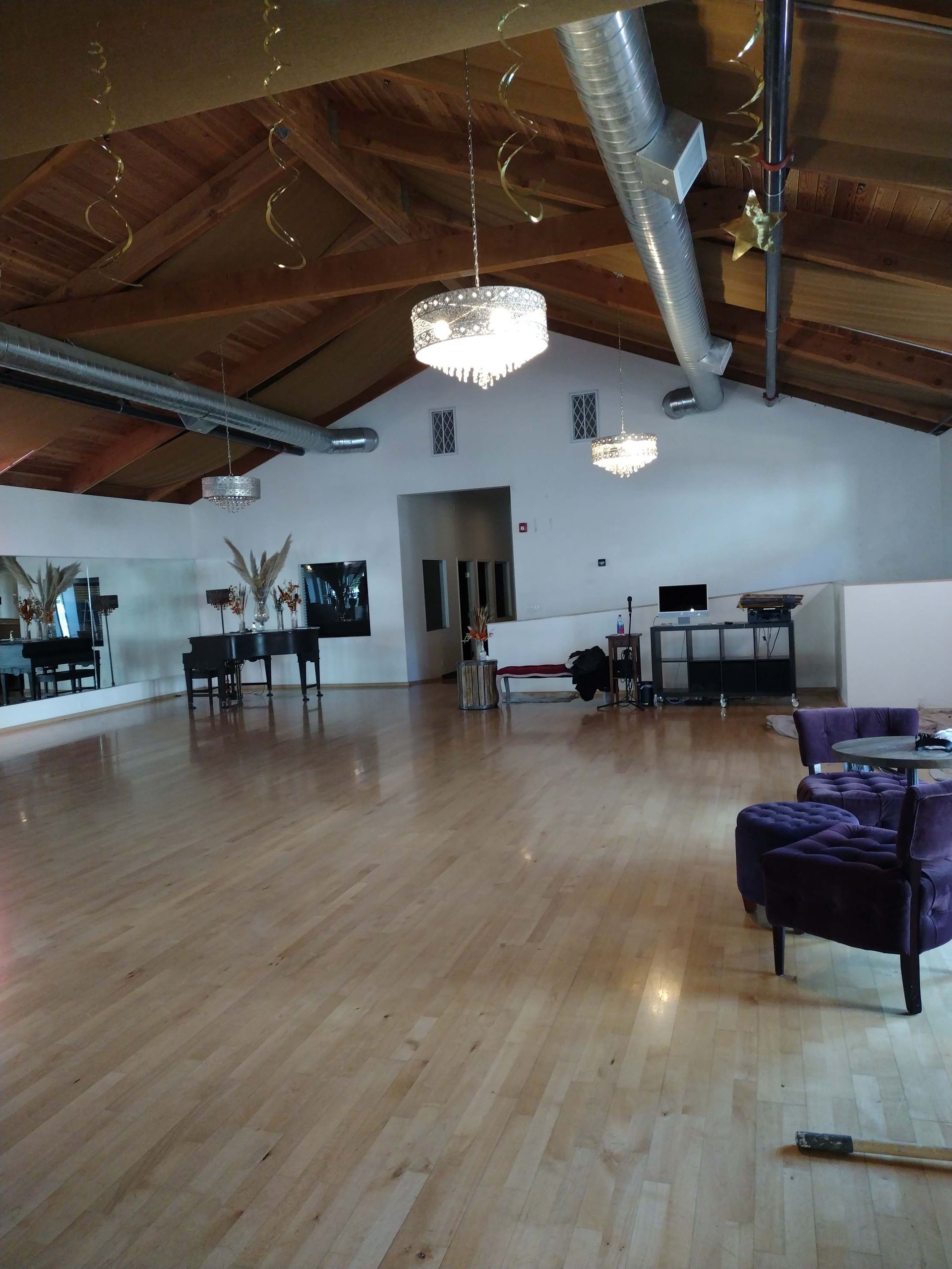 An indoor dance studio with wood floors, mirrors, vaulted wood ceilings, crystal chandeliers, and purple velvet chairs.