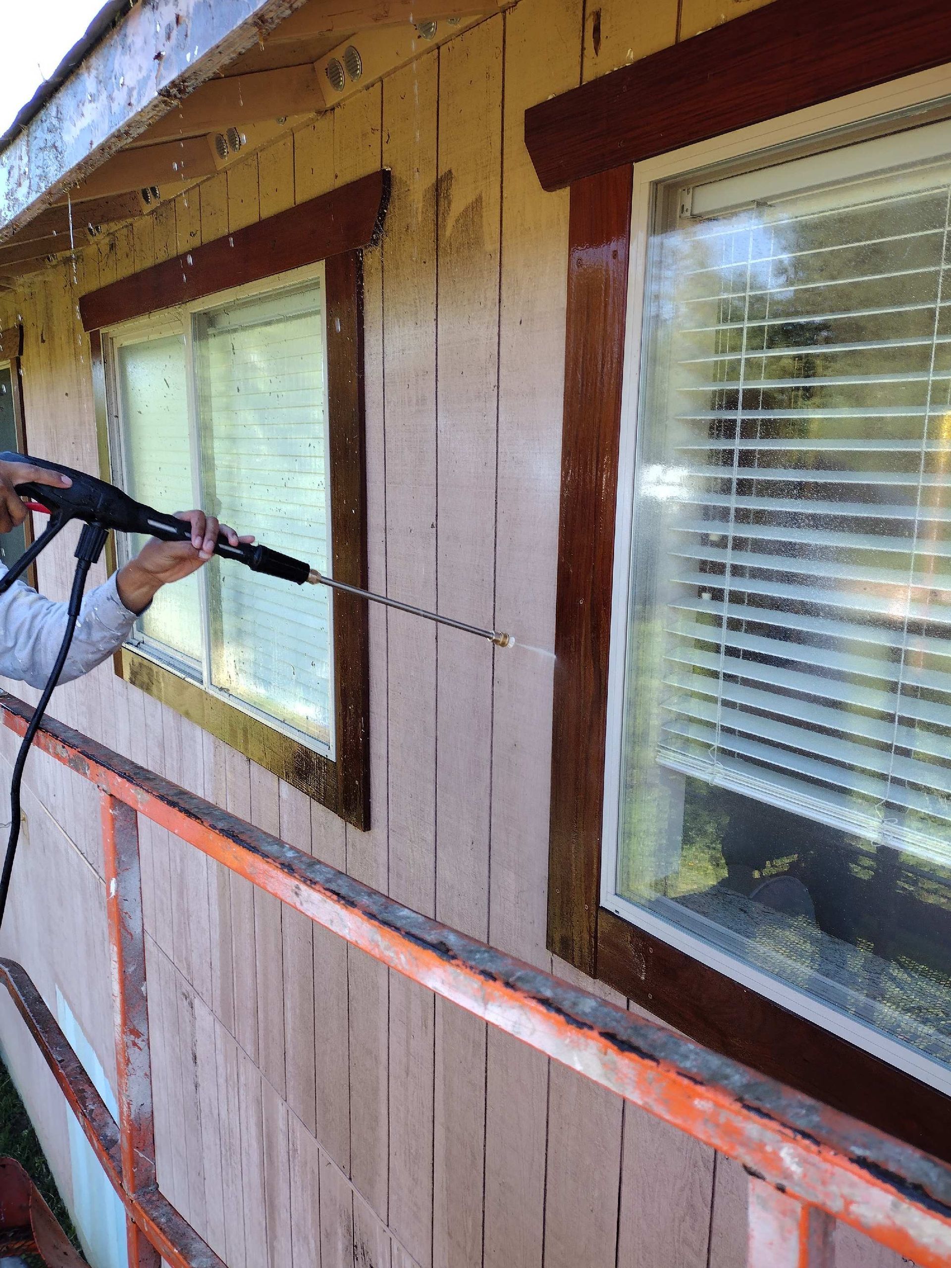 A person in a yellow raincoat and face shield uses a pressure washer to clean the light-colored siding of a house.