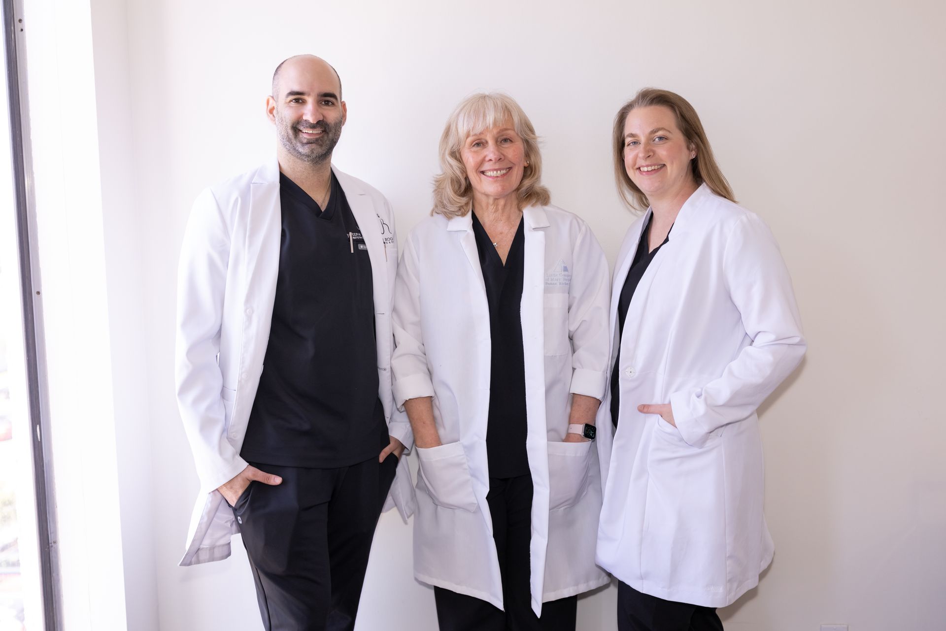 Three smiling medical professionals in white lab coats and black scrubs standing side-by-side against a white wall.