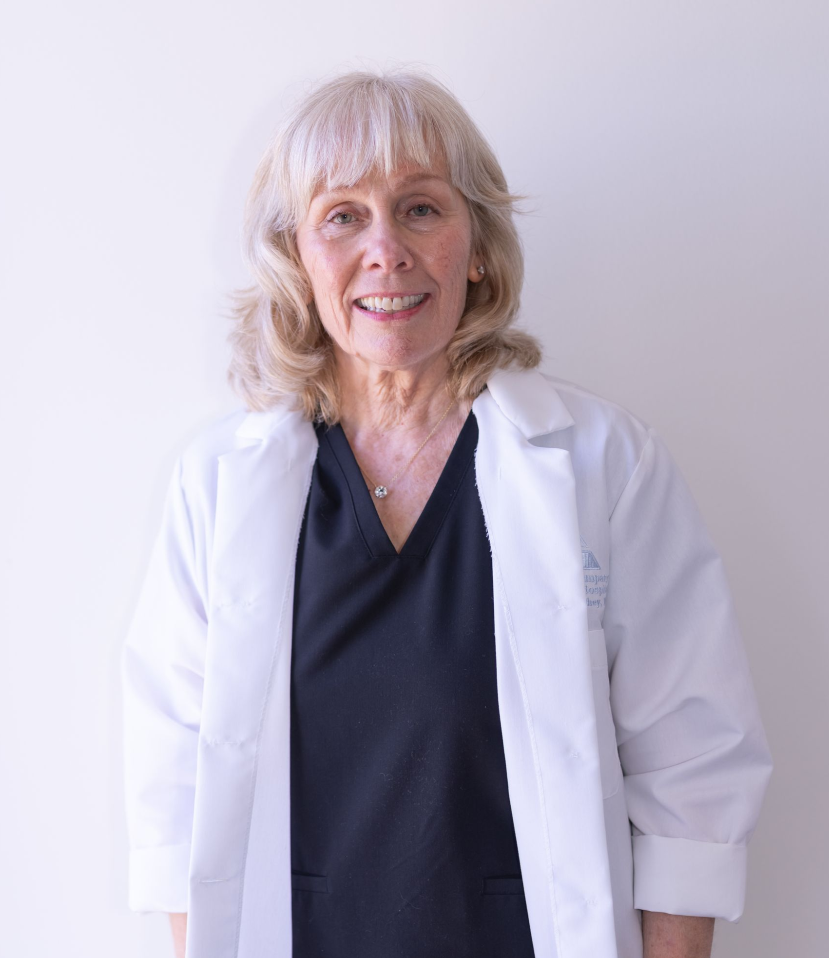 A smiling person with shoulder-length blonde hair and bangs, wearing a black and white patterned shirt and a dark pendant.