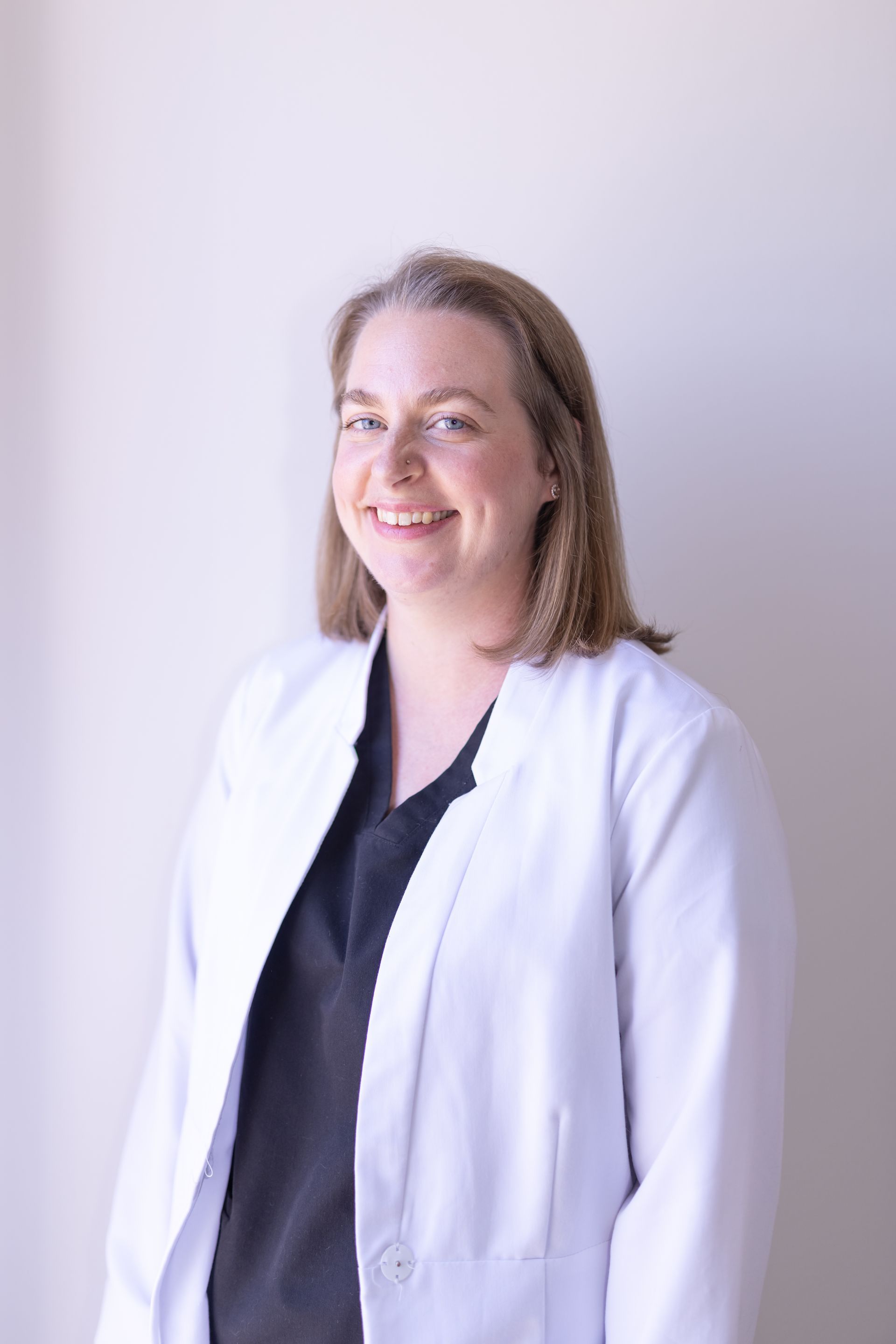 A smiling person with long blonde hair, wearing a maroon shirt and a stethoscope around their neck, sits in an office.