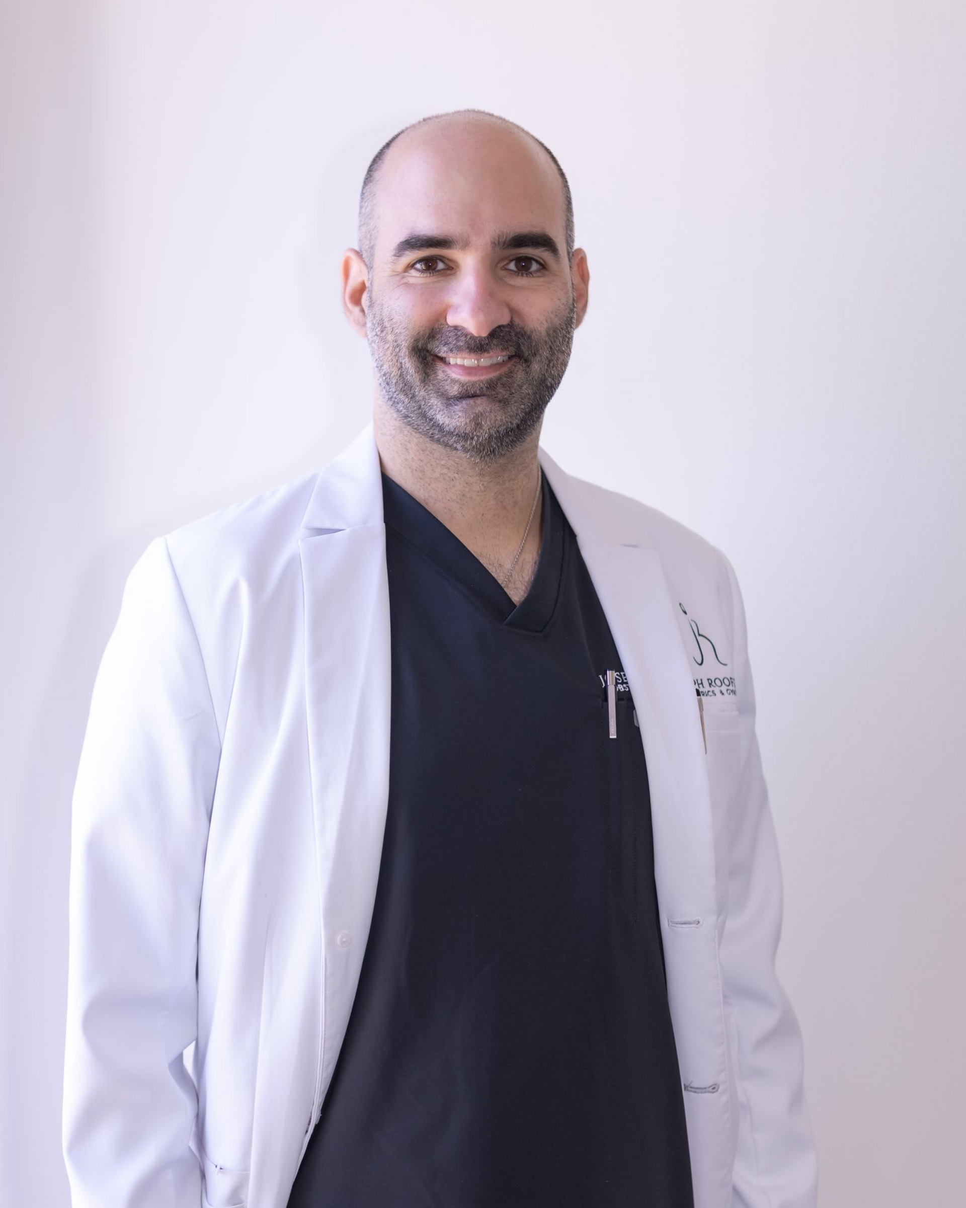 A smiling person with shoulder-length blonde hair and bangs, wearing a black and white patterned shirt and a dark pendant.