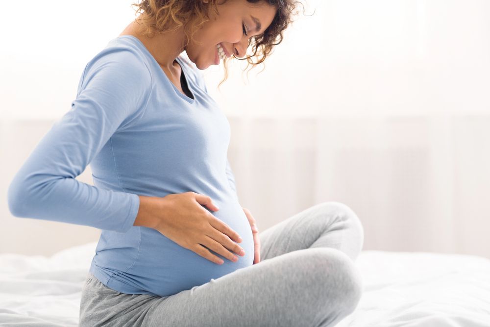 Pregnant person in a blue shirt and gray pants smiles while touching her belly; sitting on a bed.