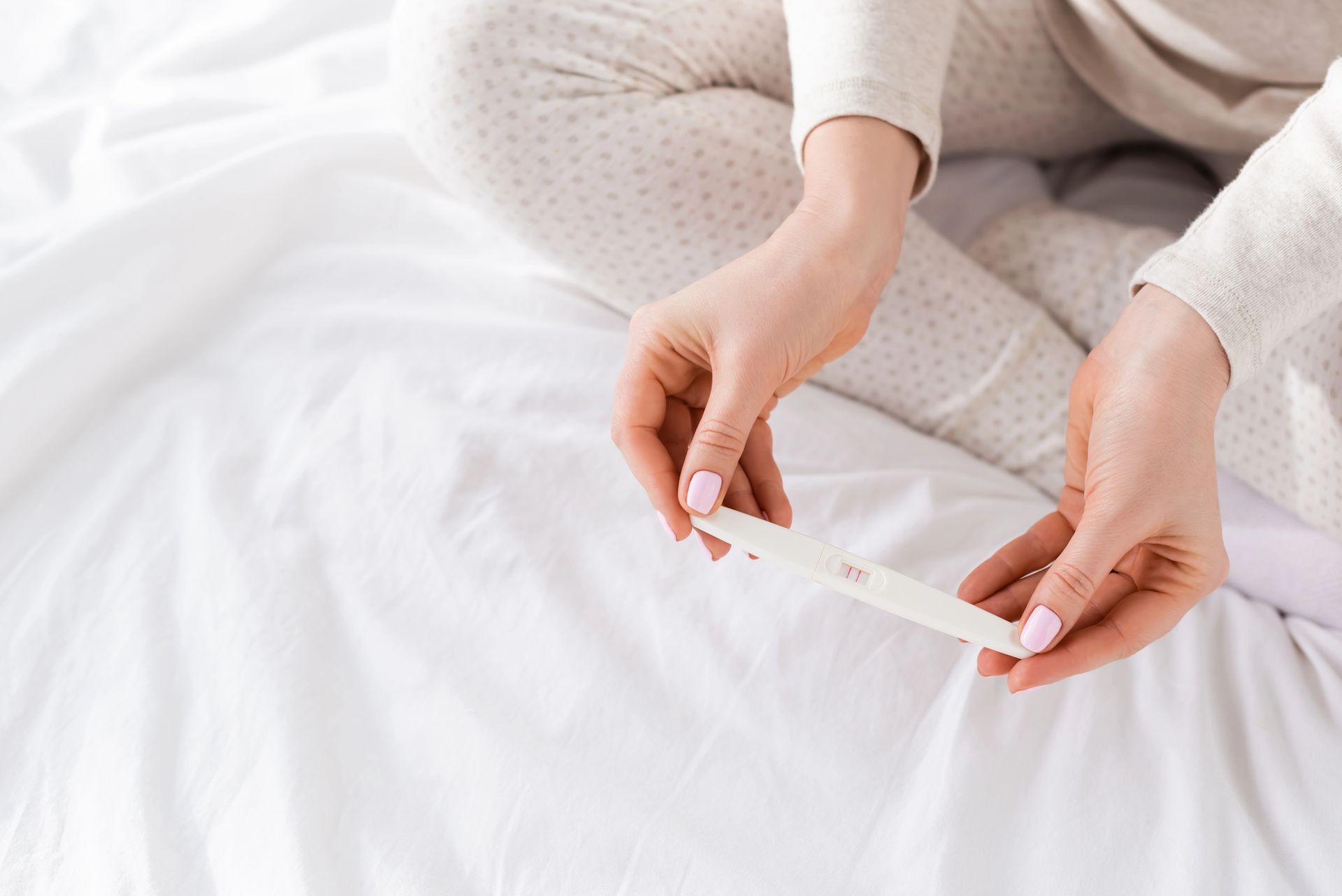 Woman in pajamas on white bed holding a positive pregnancy test.