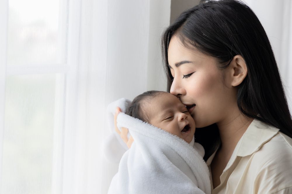 Woman kissing a sleeping baby wrapped in a white towel by a window.