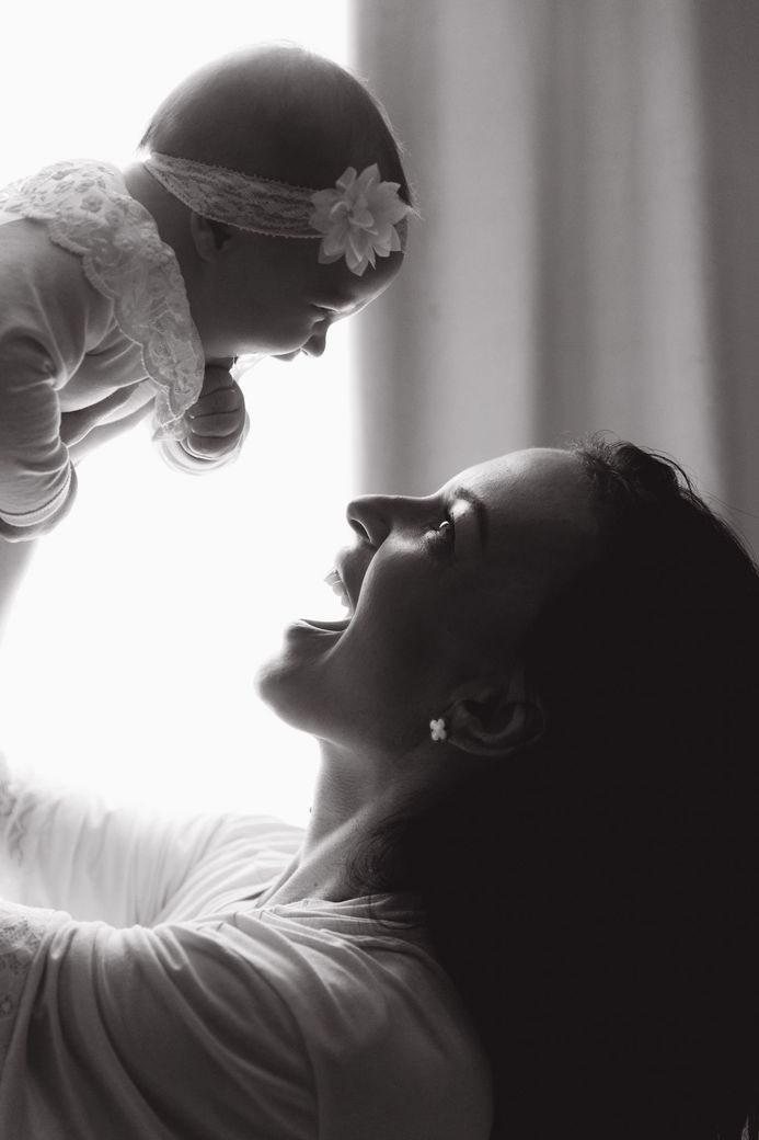 Woman holding a baby up, smiling. Baby wears a headband and lace. Black and white photo.