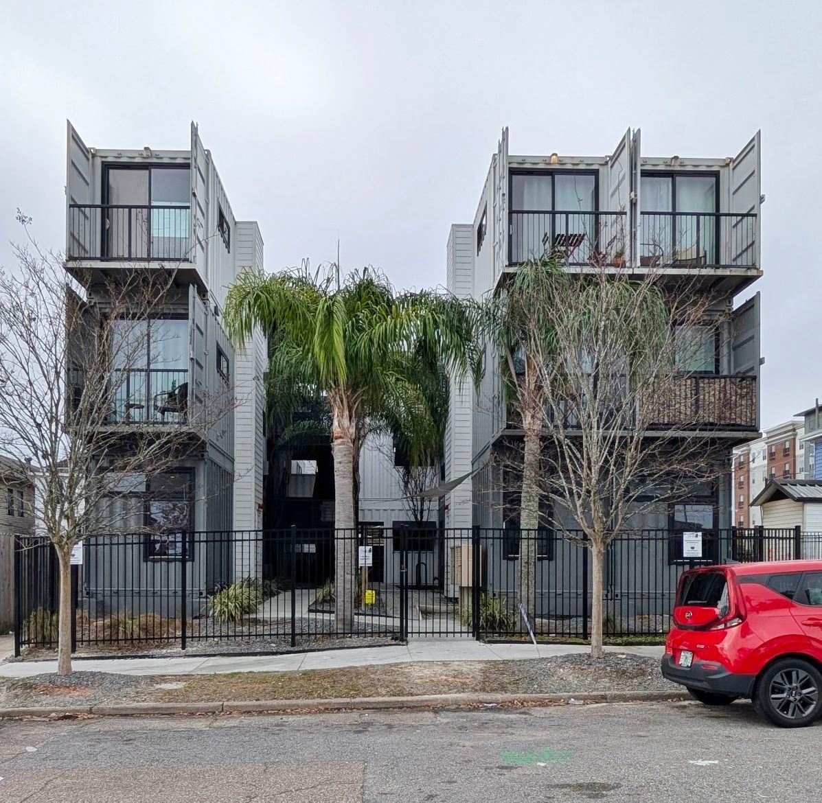 Modern gray apartment building with balconies, trees, and a red car parked on the street.