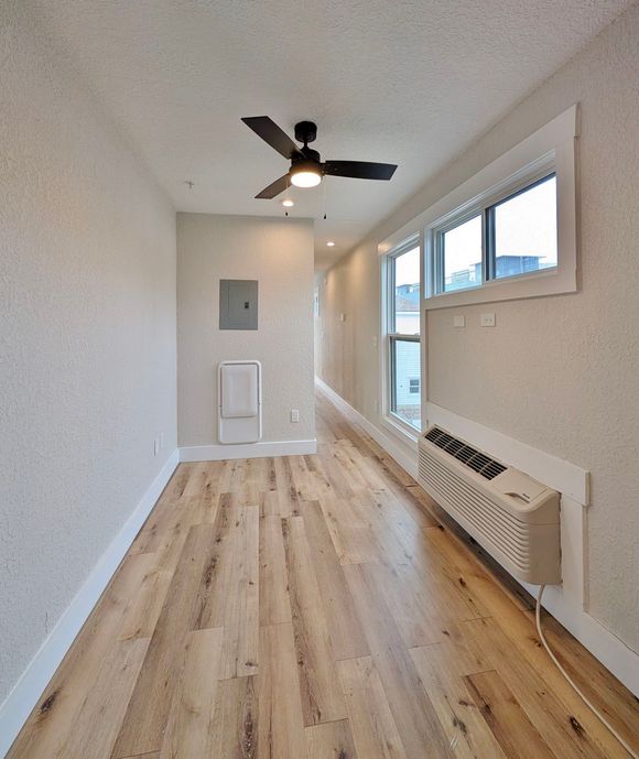 Narrow room with wood floor, white walls, ceiling fan, window, and wall heater.