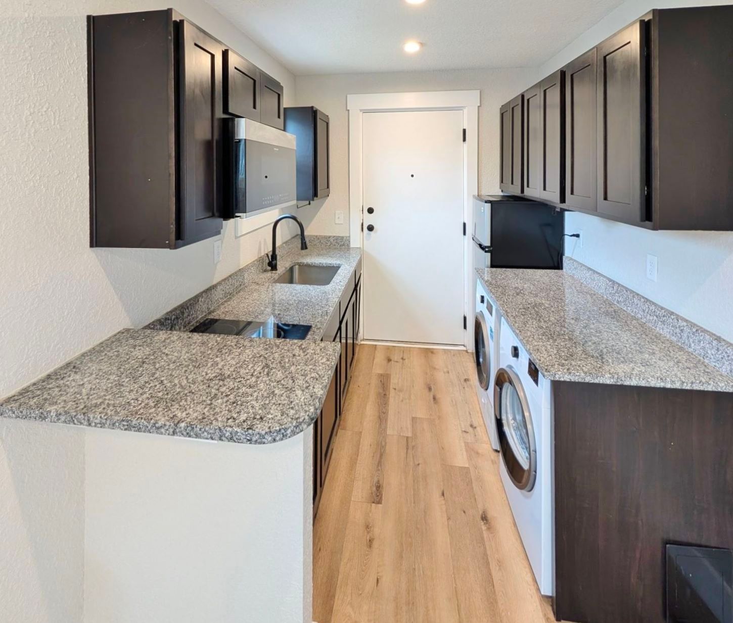 Modern galley kitchen with dark cabinets, granite counters, and light wood floor. Washing machine visible.
