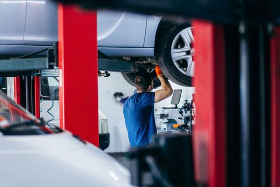 Mechanic working under a car on a lift in a shop; red and white columns, tire visible.