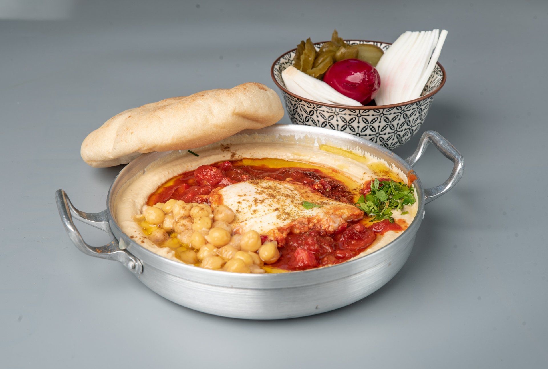 תבנית מלאה בחומוס וחומוס לצד קערת חמוצים וכיכר לחם.