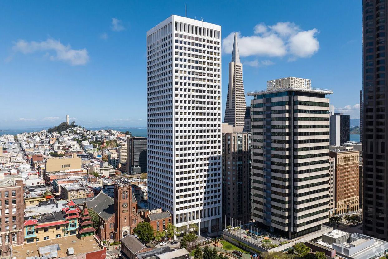 City skyline with tall office towers, dense low-rise buildings, and a distant bay under a clear blue sky