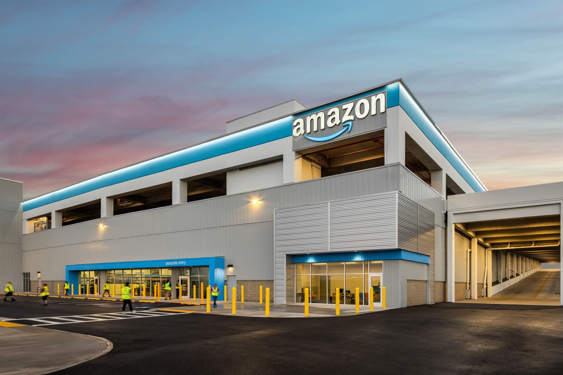 A modern Amazon facility at dusk with a large company logo, multi-level parking, and workers in high-visibility vests.