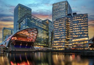 Modern glass office towers at dusk beside a curved, illuminated waterfront pavilion.