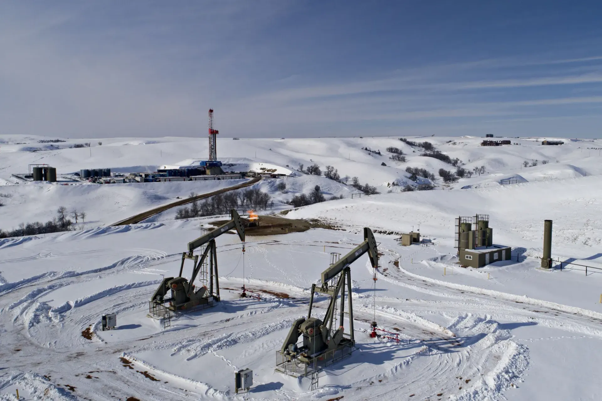 An oil drilling rig and two pump jacks operate in a snowy, hilly landscape under a clear blue sky.
