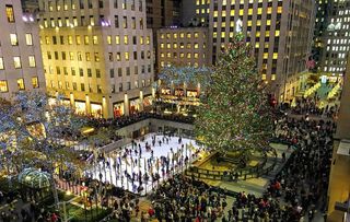 Ice skating rink in a crowded city plaza at night, surrounded by tall lit buildings and a decorated Christmas tree