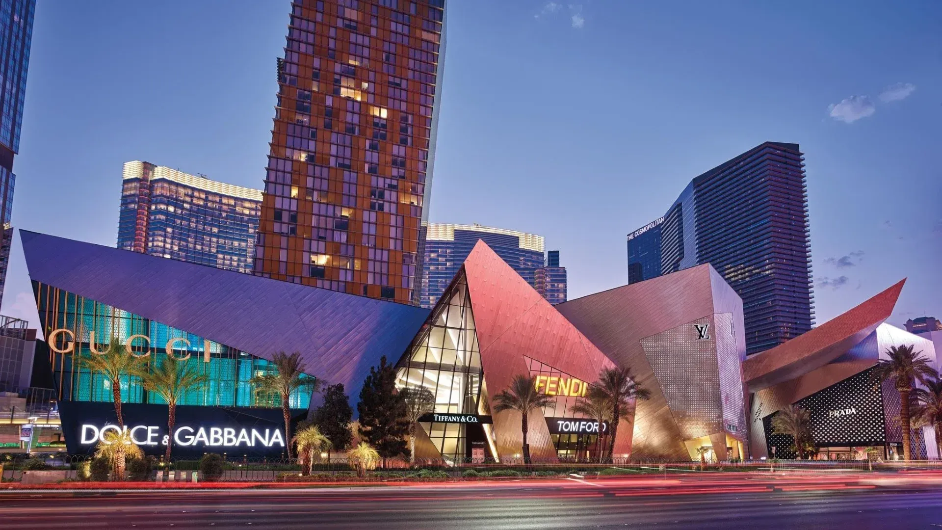 Modern shopping plaza with glass storefronts, colorful faceted facade, and tall city towers at dusk