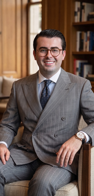 Man in a gray suit seated in a warmly lit office with bookshelves, smiling at the camera