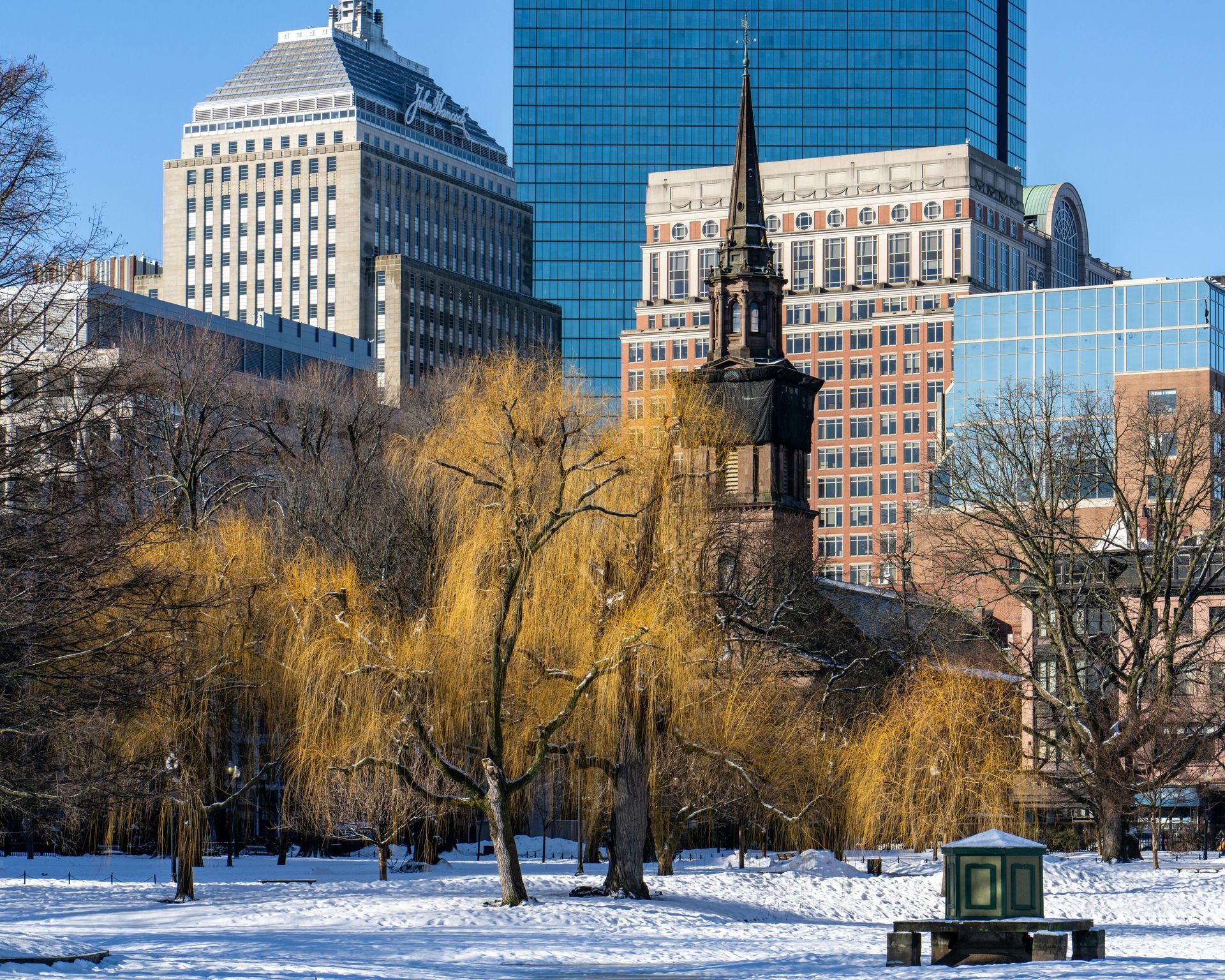 Snowy park with leafless trees and a small monument, backed by city skyscrapers in winter.