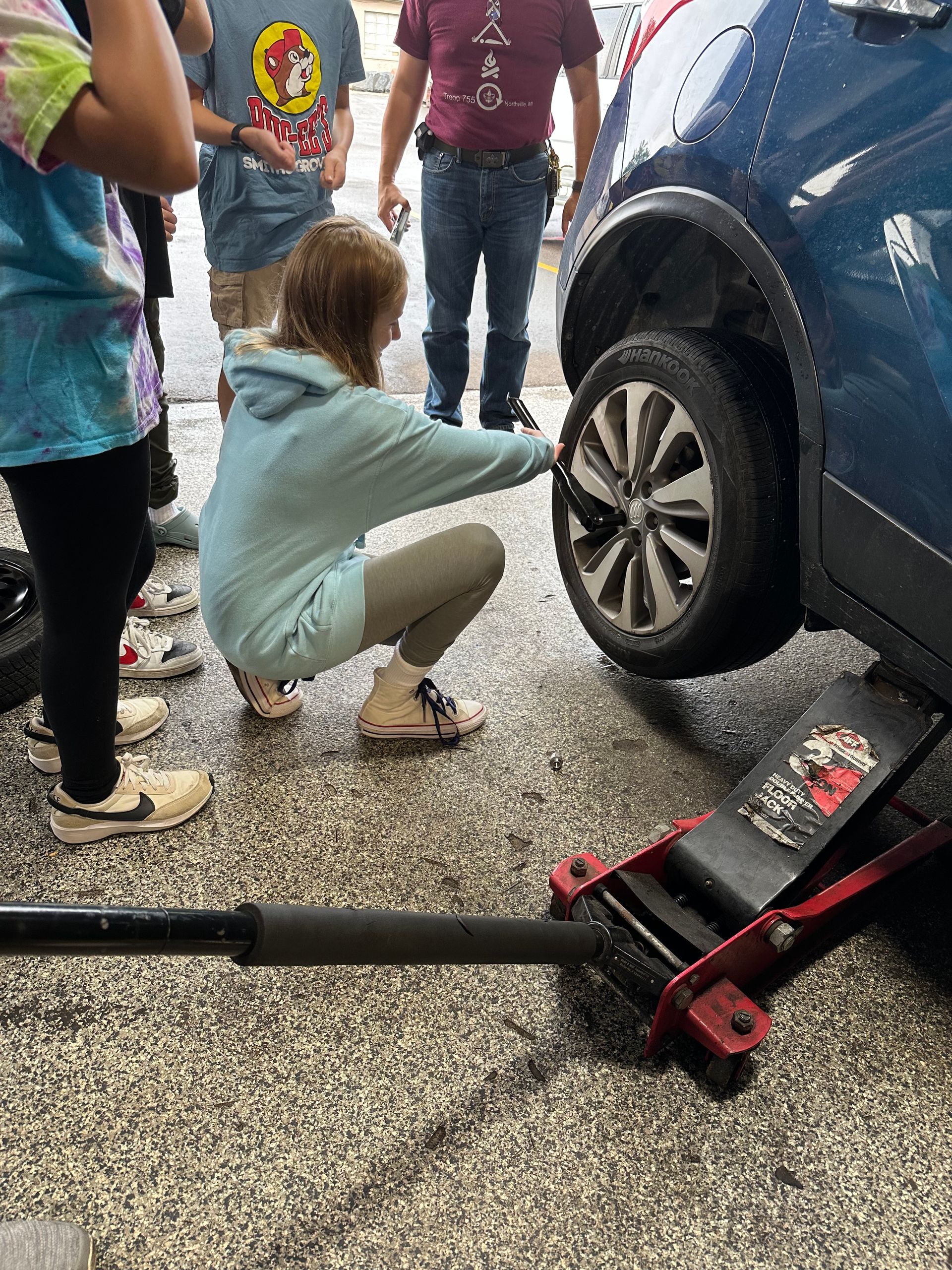 A young woman checks a car tire while squatting. Others watch in a parking area.