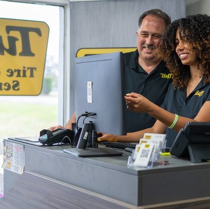 Two people working at a car tire shop counter. Woman points at computer screen, man smiles.