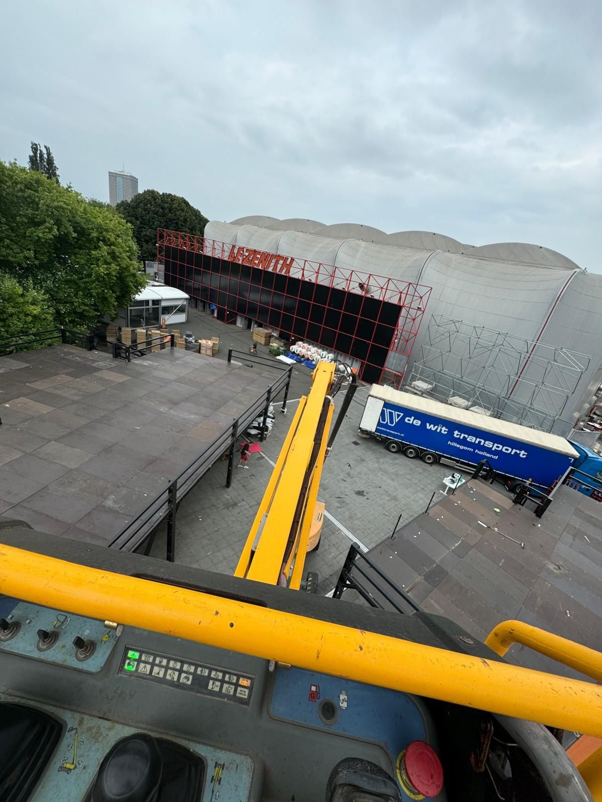 Overhead view from a yellow lift. Large building with red accents, trucks, and a loading area. Overcast sky.
