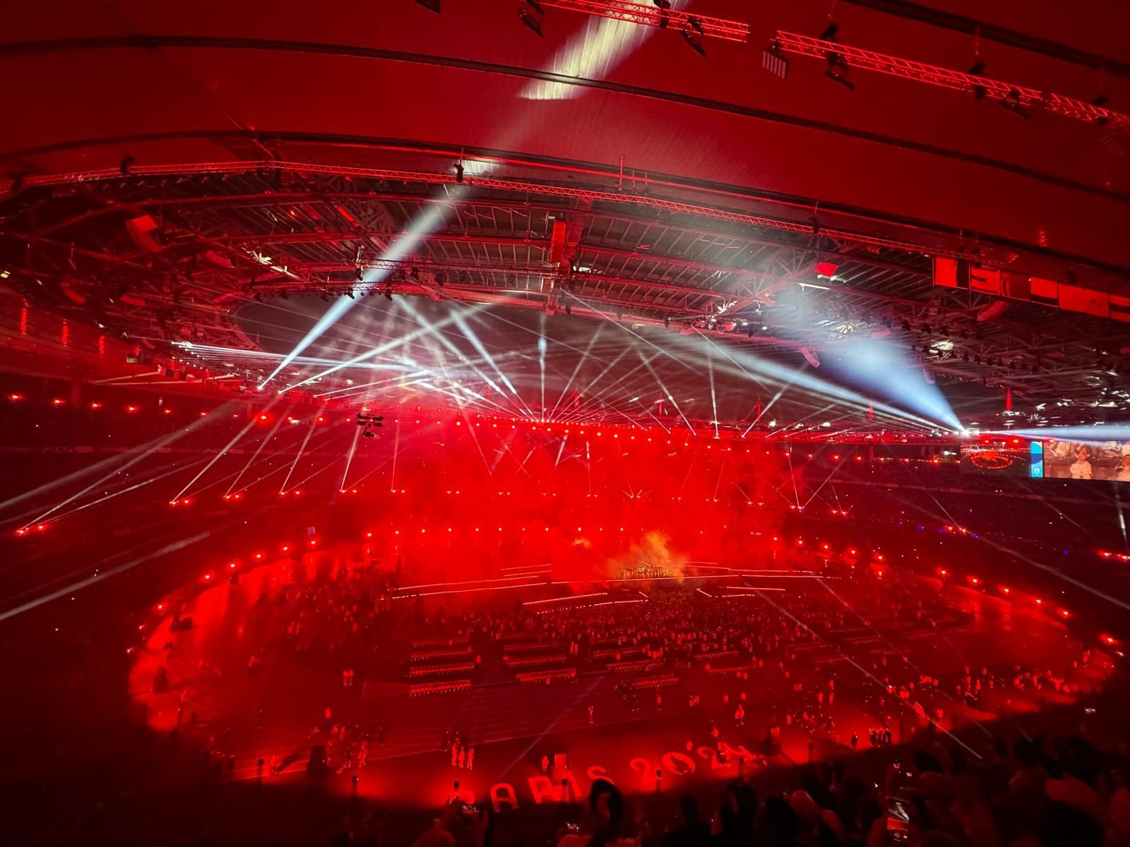 Large arena illuminated in red light with a stage and audience. Overhead spotlights beam down on a central performance area.