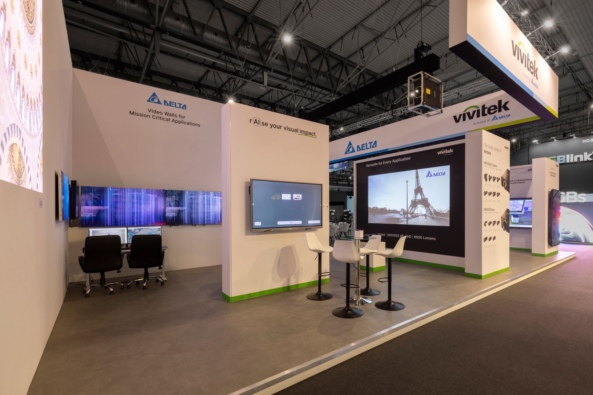 A bright trade show booth featuring Delta and Vivitek displays, wall-mounted screens, and a small table with white chairs.