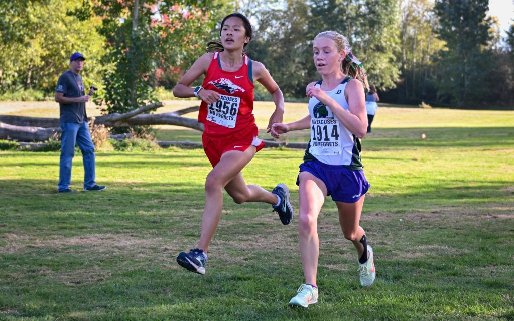 Two girls are running in a cross country race on a grassy field.