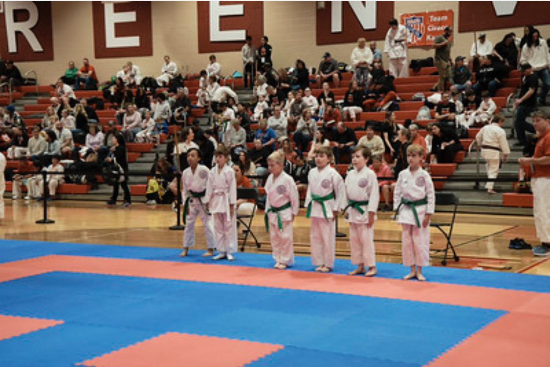 A group of kids in karate uniforms stand on a mat in front of a crowd