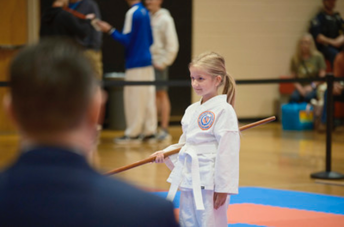 A little girl in a karate uniform is holding a stick in a gym.