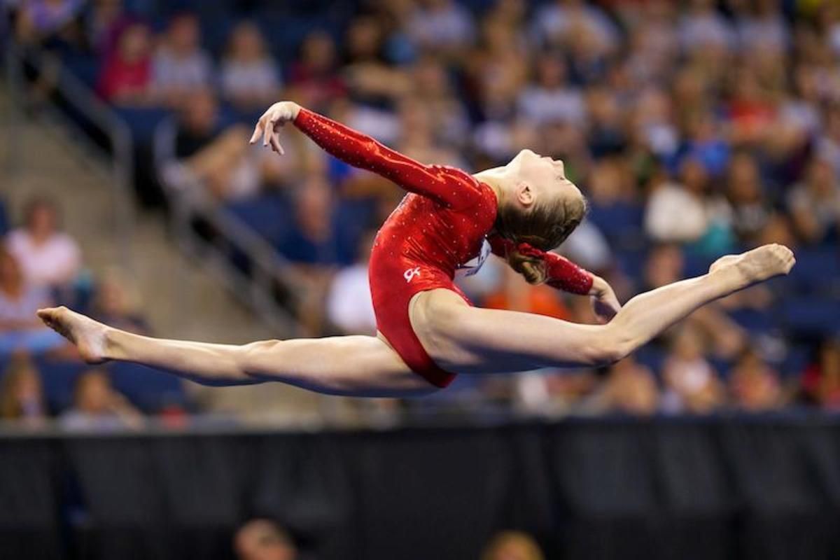 A female gymnast in a red leotard is doing a split in the air.