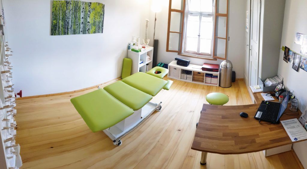 Physiotherapieraum mit Behandlungsliege, Schreibtisch und Fenster. Grün-weiße Einrichtung, Holzboden.